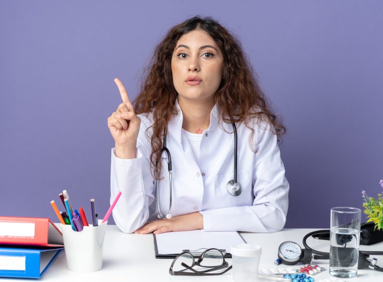 impressed-young-female-doctor-wearing-medical-robe-stethoscope-sitting-table-with-medical-tools-looking-front-pointing-up-isolated-purple-wall